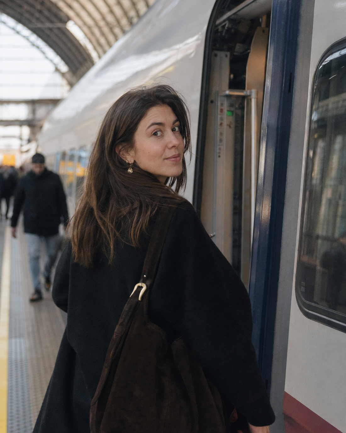 A woman glancing back over her shoulder while boarding a train at a glass-roofed station platform, wearing a small gold drop earring and carrying a dark suede tote bag