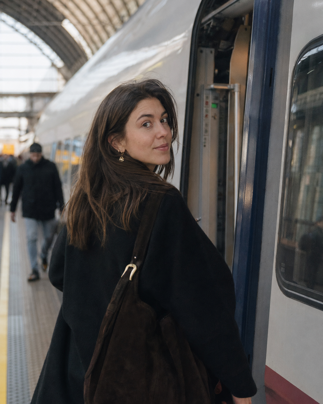 A woman glancing back over her shoulder while boarding a train at a glass-roofed station platform, wearing a small gold drop earring and carrying a dark suede tote bag