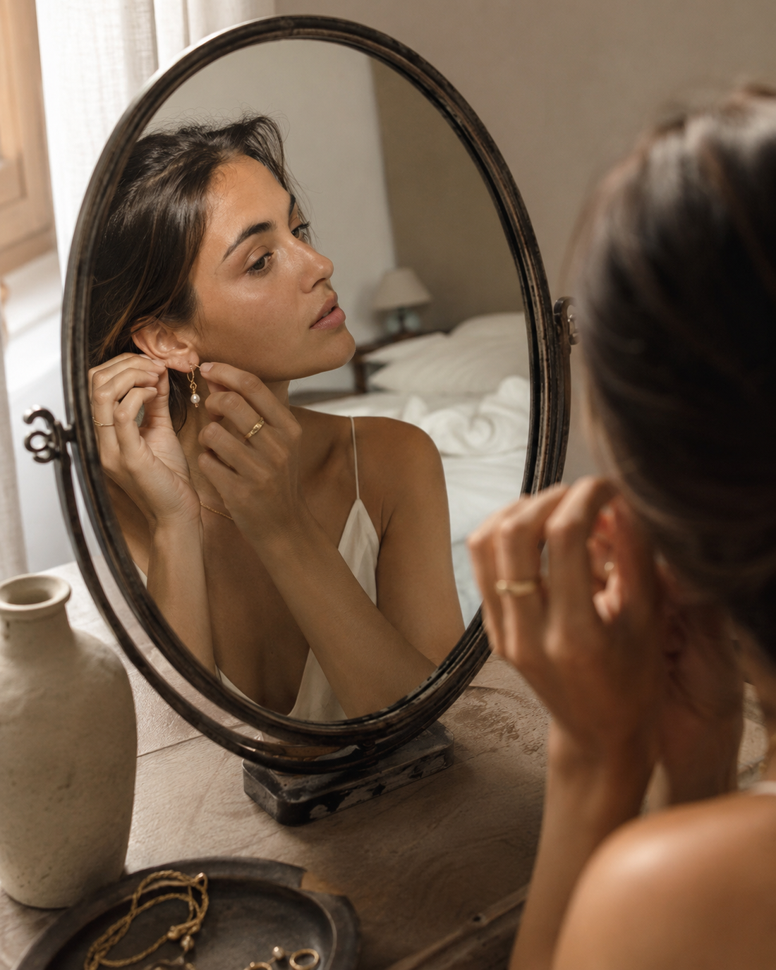 A woman leans toward an antique oval mirror, gently fastening a small gold earring, with a shallow dish of jewelry visible on the vanity beside her