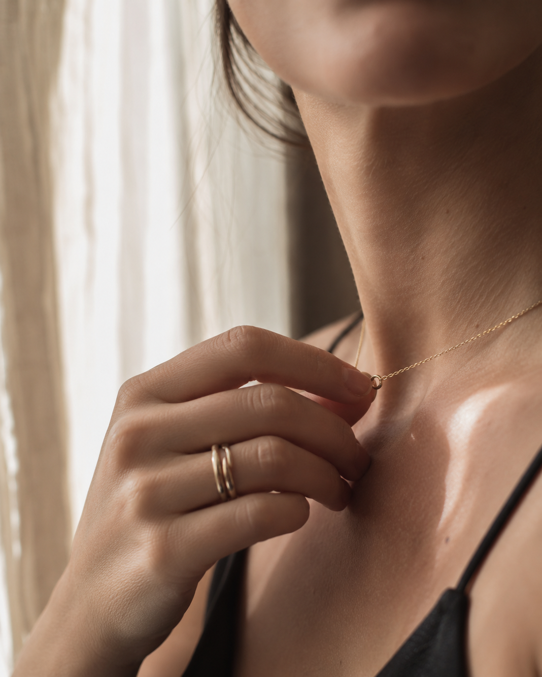 A woman's hand, two thin gold rings on her fingers, gently touching a fine gold necklace at her collarbone in warm afternoon light