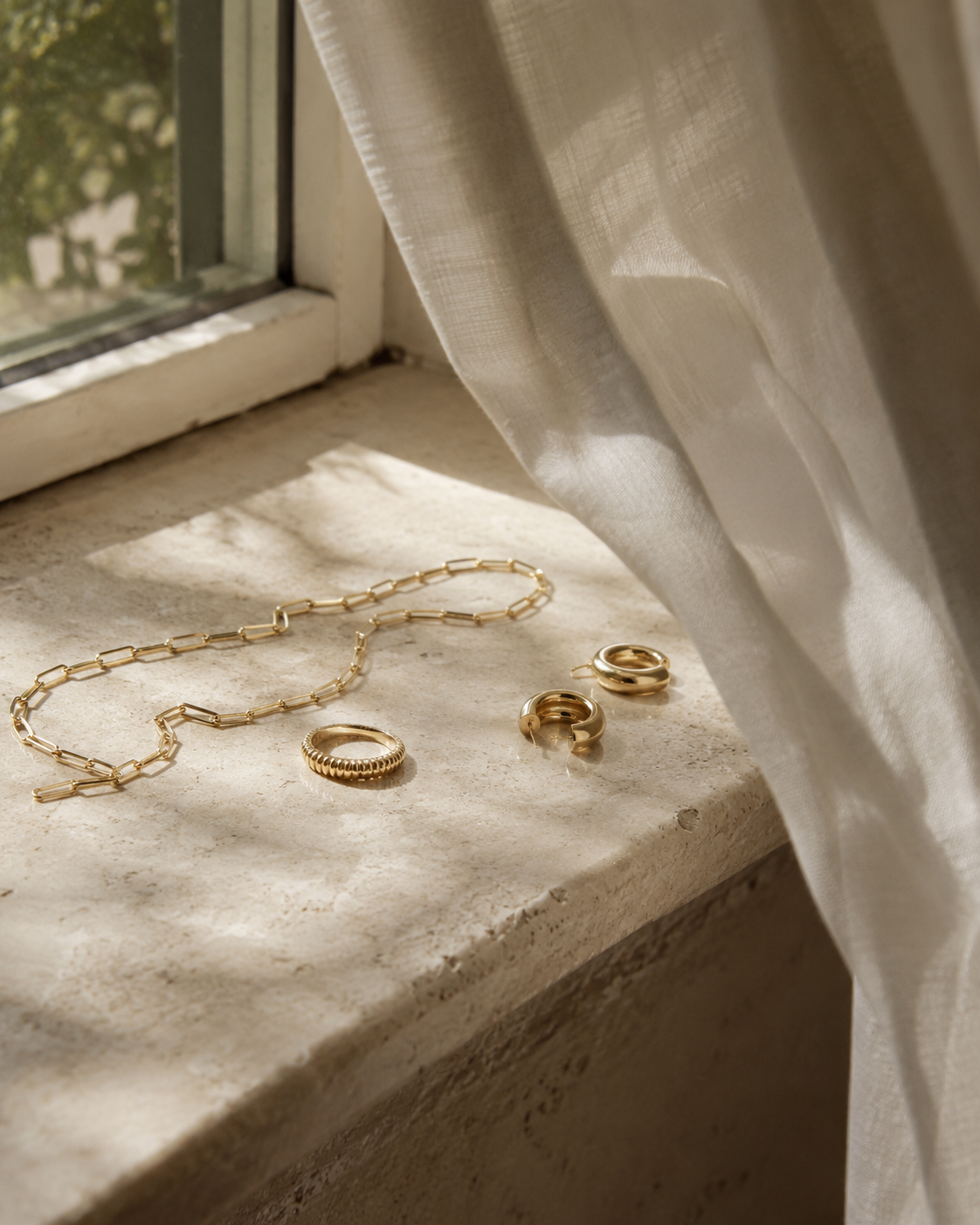 A gold paperclip chain necklace, a textured ring, and two small gold hoop earrings laid on a sunlit stone windowsill beside a softly billowing linen curtain