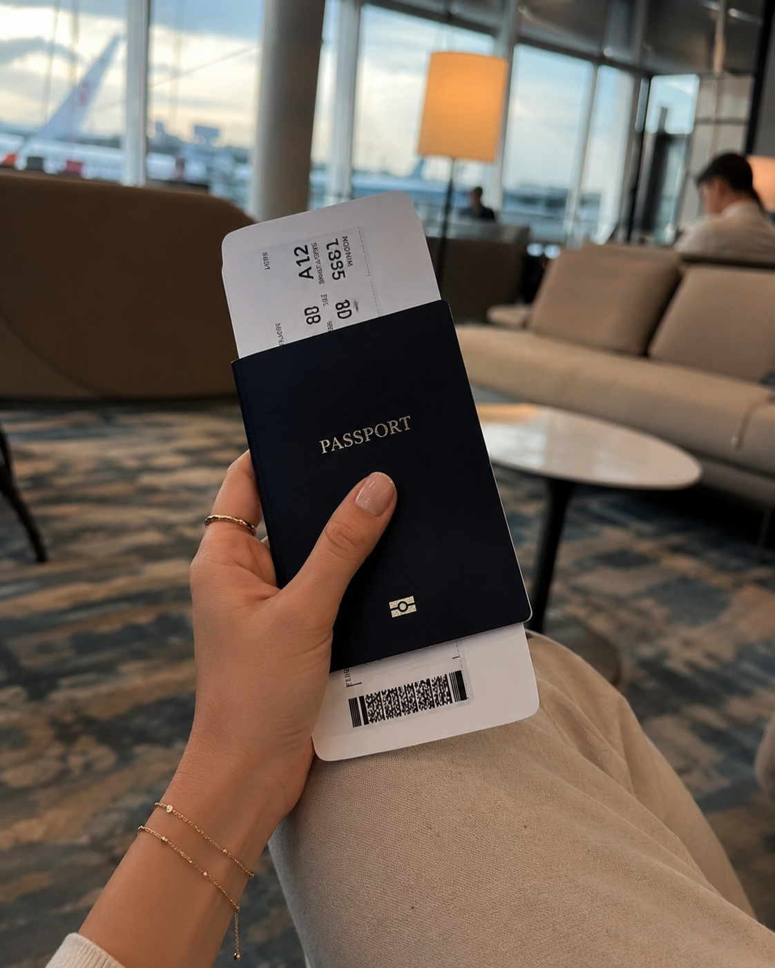 A hand adorned with thin gold rings and a delicate chain bracelet holds a navy passport and boarding pass in an airport lounge at dusk, with a glowing lamp and an airplane visible through the window behind.