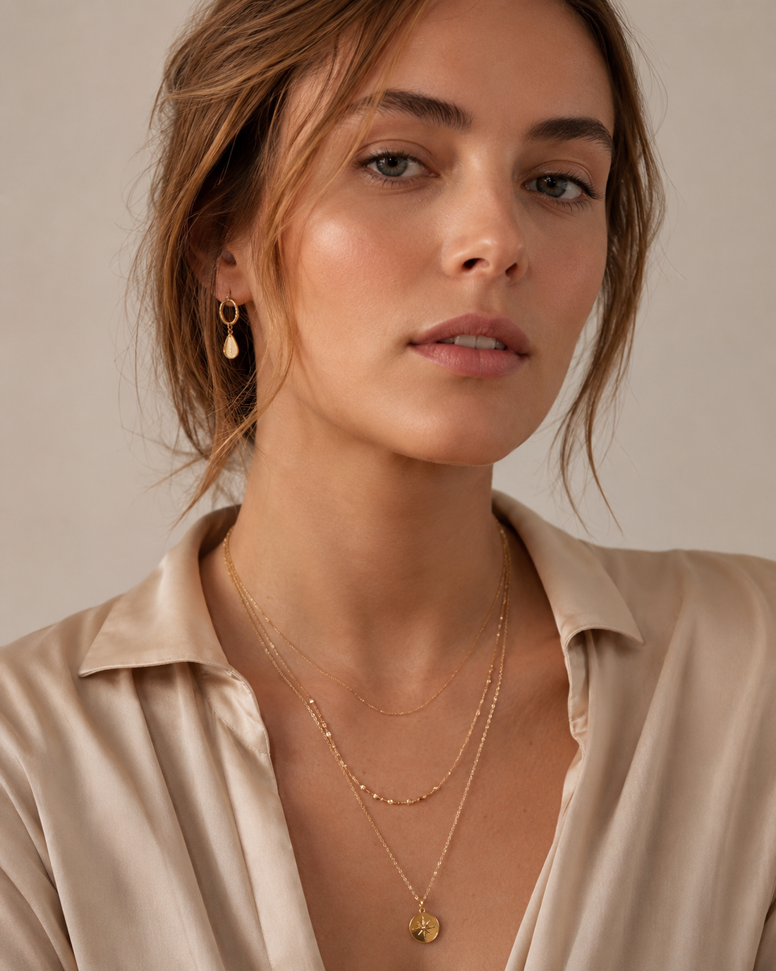 A close-up portrait of a woman wearing layered thin gold necklaces and a gold pearl-drop earring against a soft neutral backdrop.