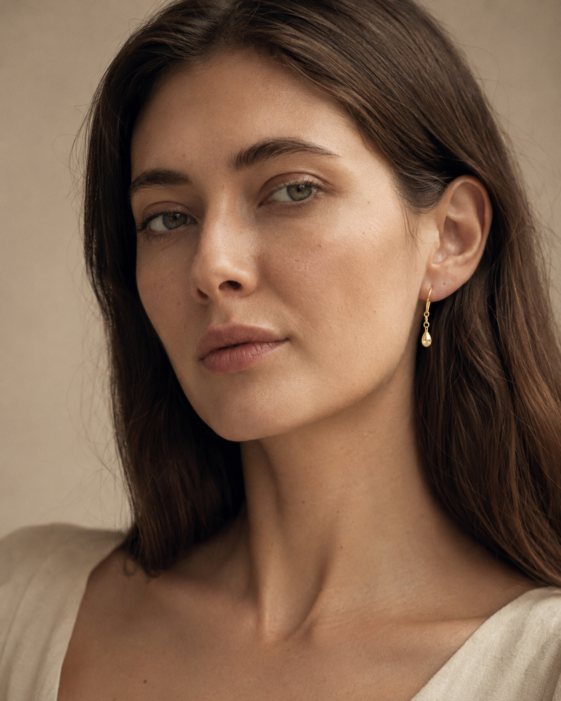 A close-up studio portrait of a woman with dark hair, wearing a delicate gold drop earring, softly lit against a warm neutral background.